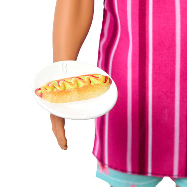 Close-up of Ken doll's hand holding a plate with a hotdog play piece, with the pink striped apron