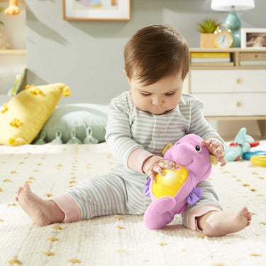 Baby sitting on a rug squeezing the pink Fisher-Price Soothe & Glow Seahorse